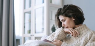 Zamyślona kobieta w swetrze patrzy w smartfon, siedząc samotnie na kanapie